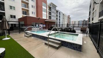A swimming pool with steps leading into it, surrounded by a fence, and adjacent to a building with multiple balconies.