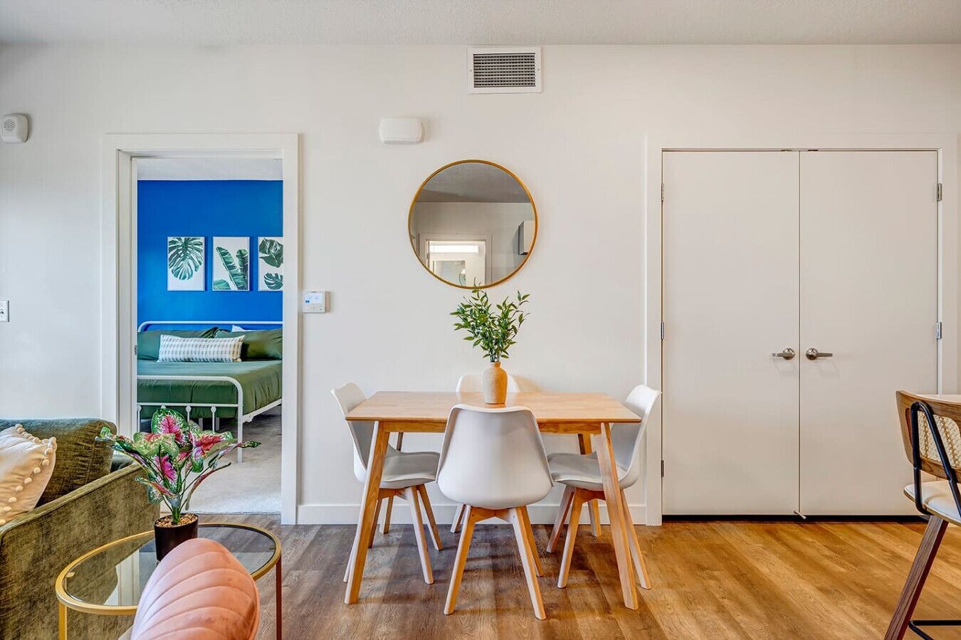 A dining area with a wooden table and chairs, a round mirror, and a potted plant.