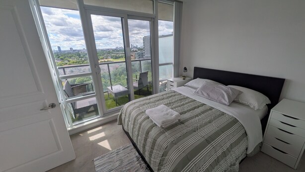 Ein Schlafzimmer mit großem Fenster, einem Bett mit gestreifter Bettwäsche, einem Nachttisch und einem Stuhl vor dem Fenster.