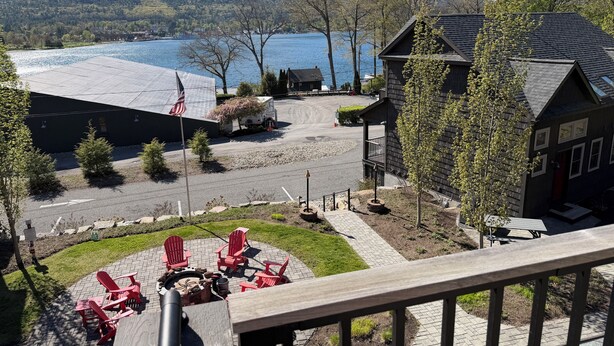 A lakeside view with a wooden deck, red Adirondack chairs, and a building with a dark roof.
