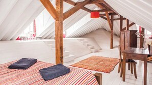Een zolderkamer met schuine wanden, houten balken, een klein tafeltje en twee bedden met kussens.