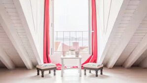 Een kamer met een hoge plafond, een tafeltje, twee stoelen en uitzicht door een raam met rode gordijnen.