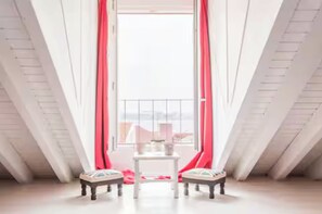 Ein Zimmer mit hohen Decken, einem kleinen Tisch, zwei Stühlen und Blick durch ein Fenster mit roten Vorhängen.