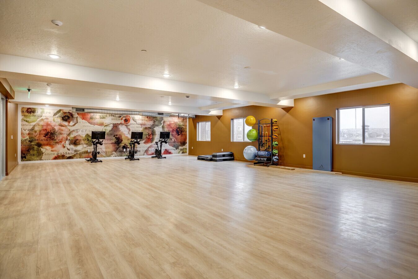 A spacious room with a wooden floor, exercise equipment, a mural, and windows.