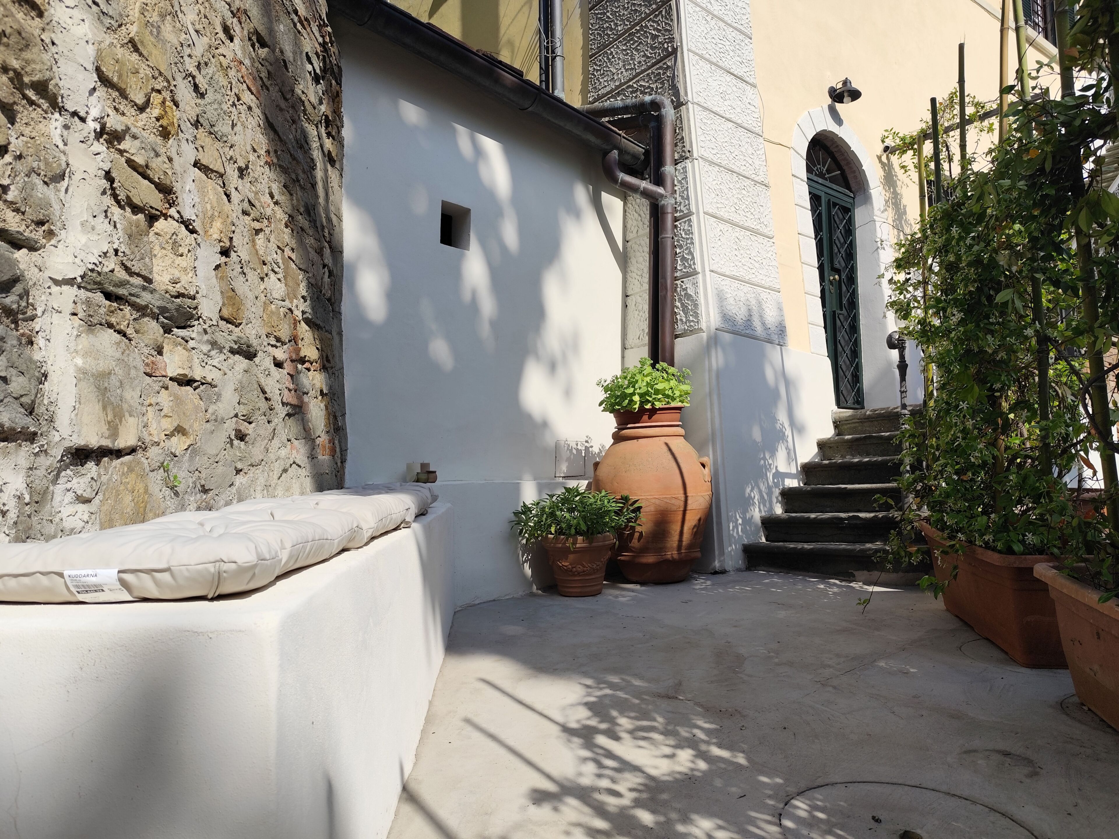 Un cortile stretto con un muro in pietra, un edificio bianco e una scala che conduce a un ingresso.