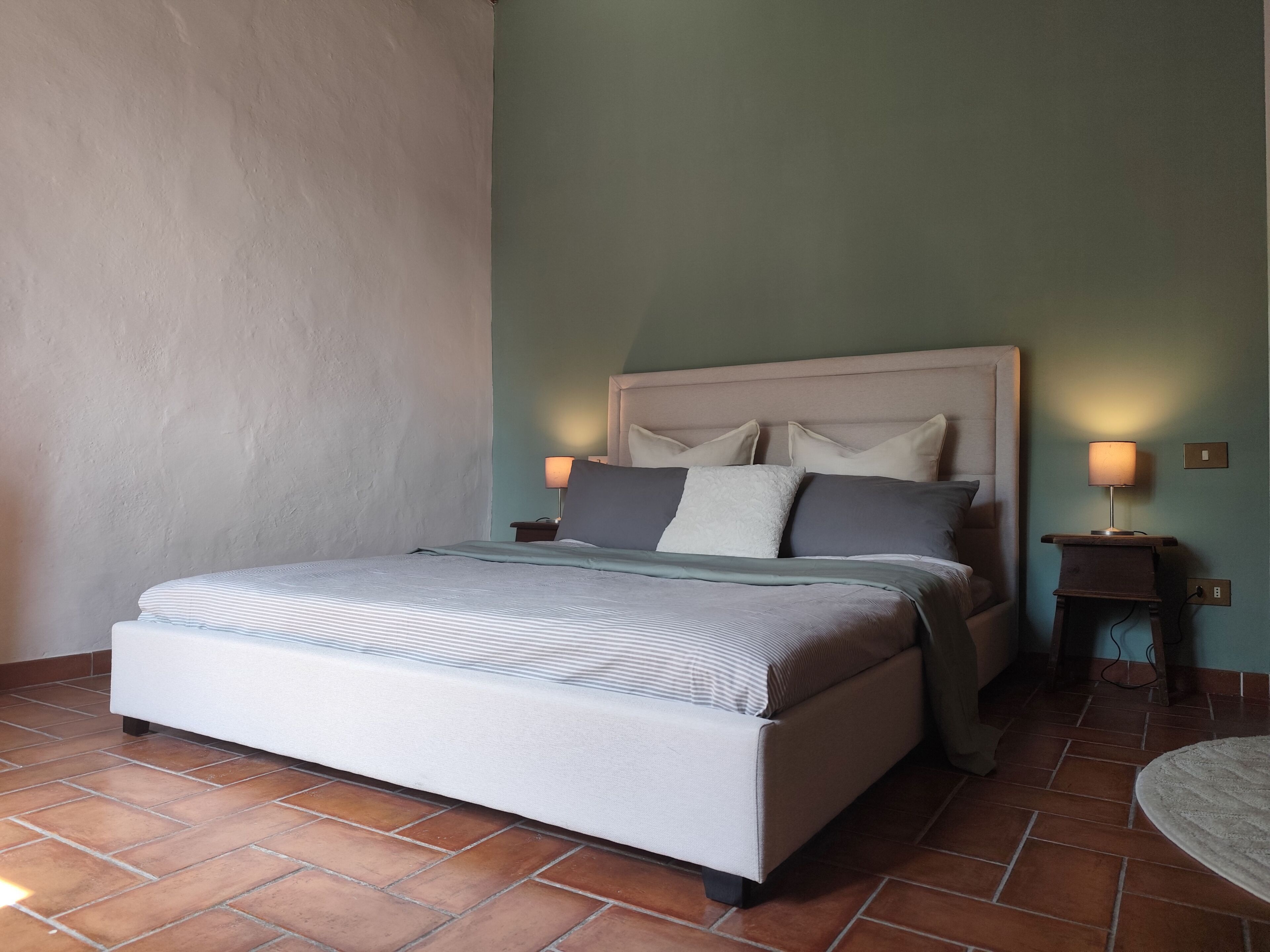 Una cama bien hecha con cabecera, dos mesitas de noche con lámparas y suelo de baldosas.