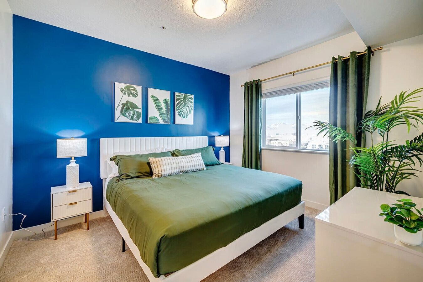 A bedroom with a bed, bedside tables, a window with curtains, and a view of the city.