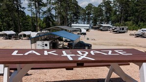 Une table de pique-nique sur laquelle est inscrit le mot « RIVE », entourée de camping-cars et située près d’un espace abrité dans un terrain de camping.