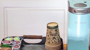 A coffee maker, a stack of disposable cups, and a container of coffee pods on a countertop.