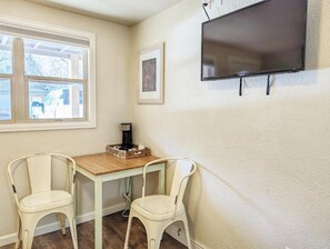 A room with a wooden table, two white chairs, a television mounted on the wall, and a framed picture. - The Fountain Creek Inn (Manitou Springs)
