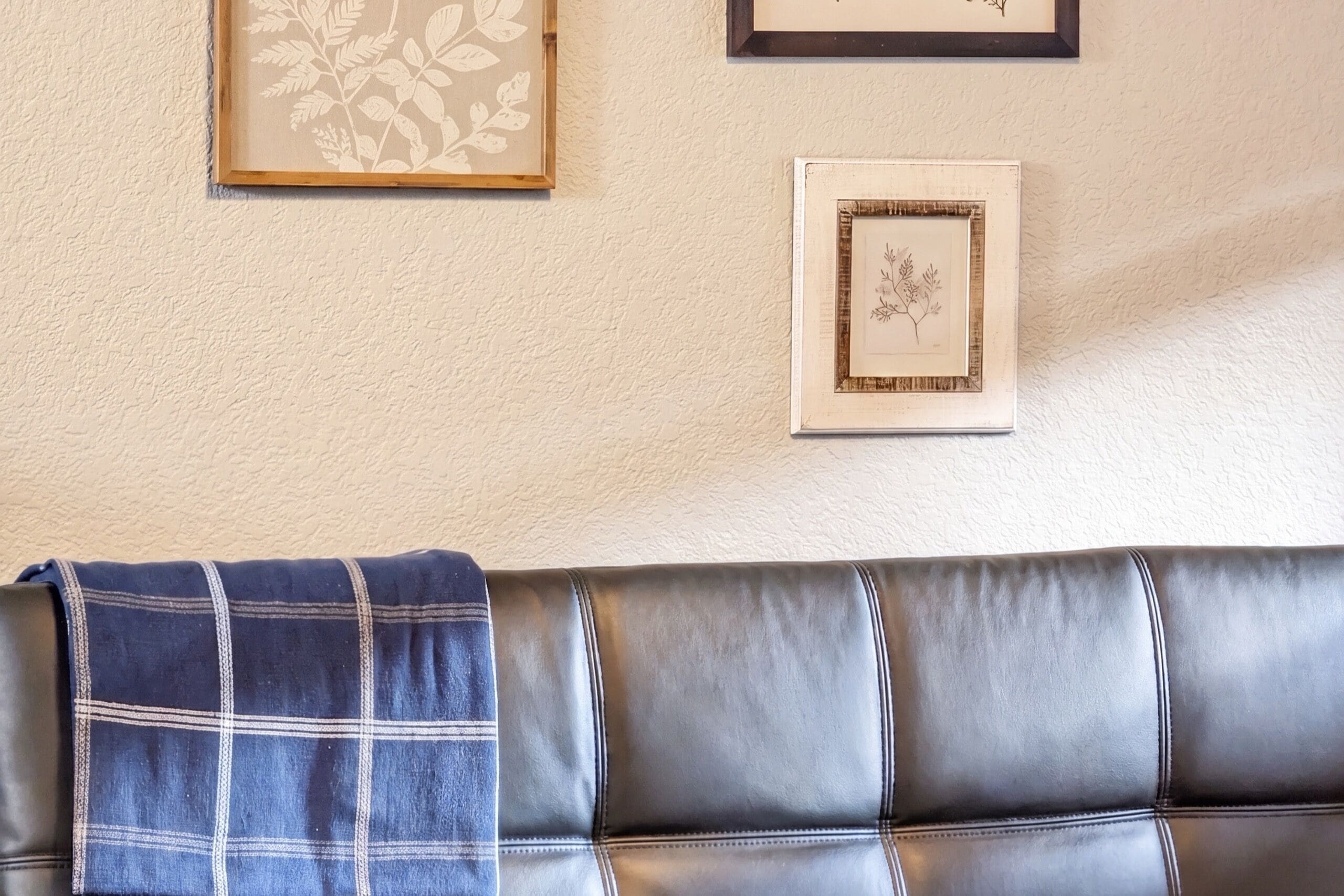 A room with a leather couch, a blue plaid throw, and two framed botanical prints on the wall.