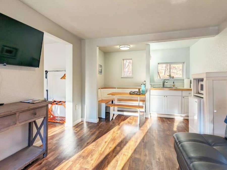 A compact living space with a wooden floor, a dining area with benches, a kitchen with white cabinets, and a TV mounted on the wall.