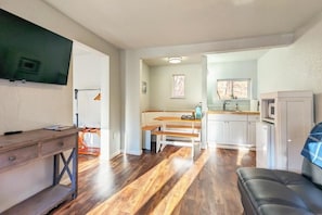 Een ruimtelijk ingerichte woonruimte met houten vloer, een eethoek met banken, een keuken met witte kasten, en een aan de muur bevestigde televisie.