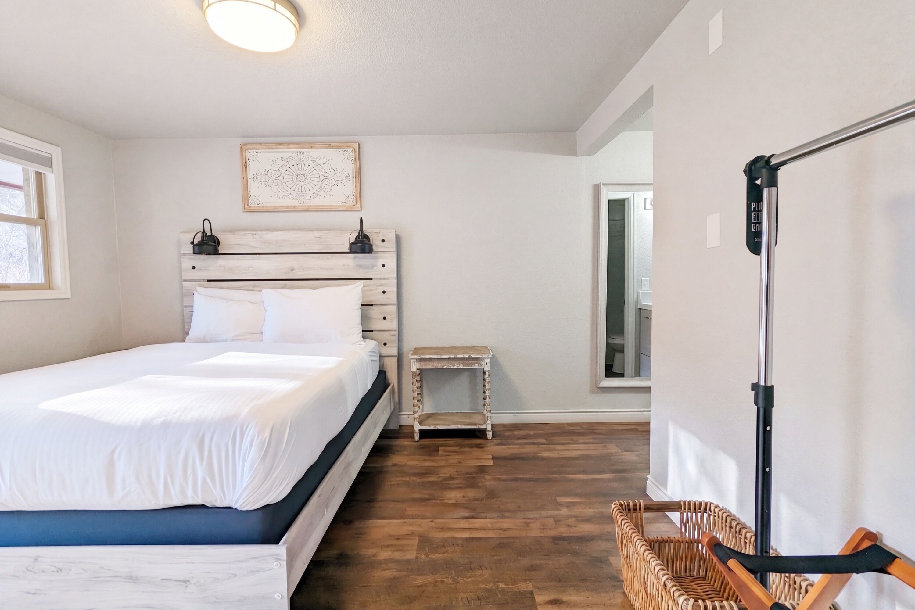 A bedroom with a bed, a bedside table, a small stool, a wicker basket, and a wall-mounted light fixture.