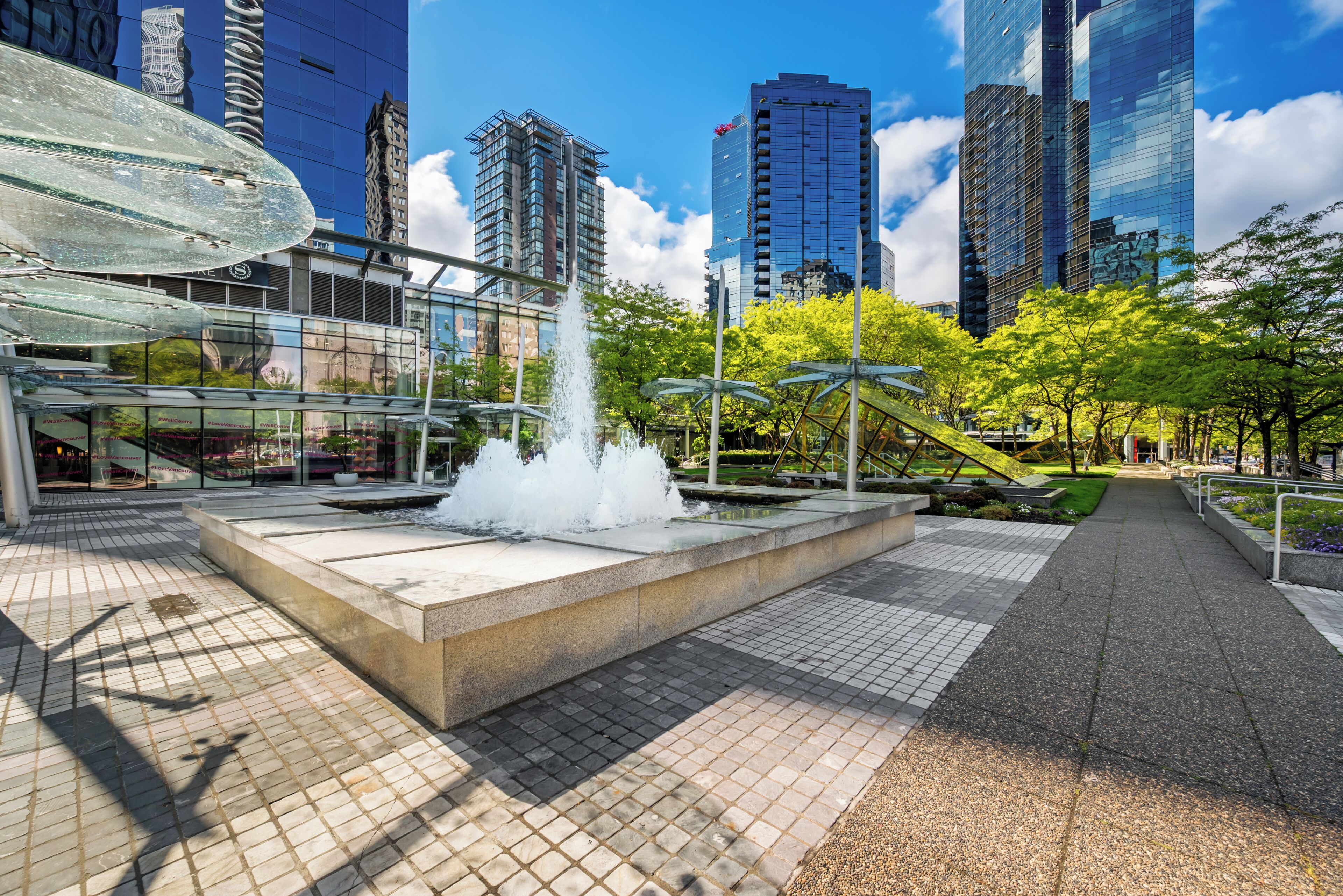 Una plaza urbana moderna con un elemento central de agua, rodeada de rascacielos y vegetación.