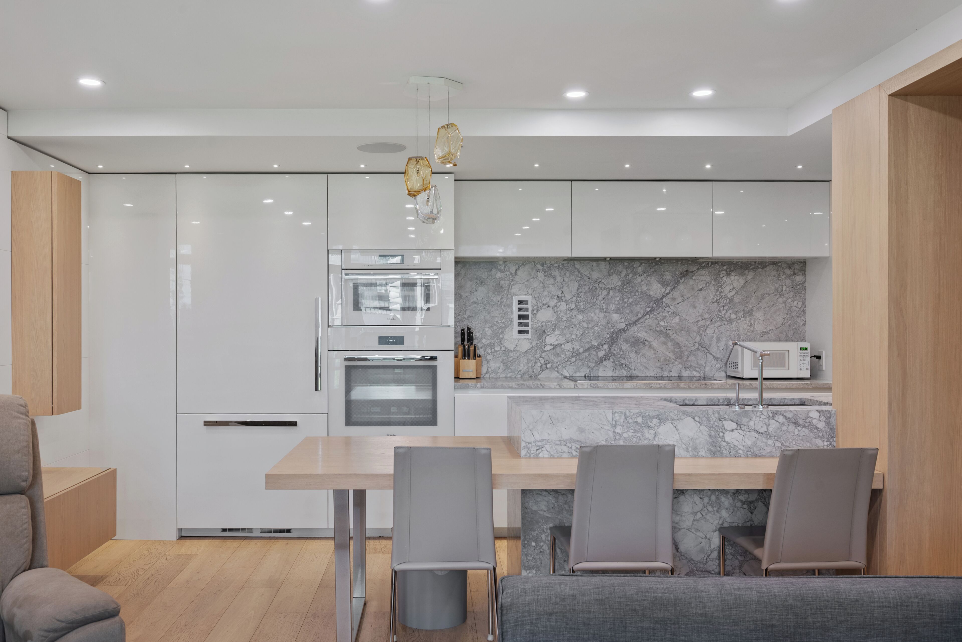 A modern kitchen with white cabinetry, a marble backsplash, and a dining area with grey chairs.