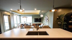 A modern kitchen with a large island, wooden cabinets, pendant lighting, and a dining area with chairs and a table. - Fernwood - Views ULTRA Comfort A+ Design 1335sf 4b (Canmore)