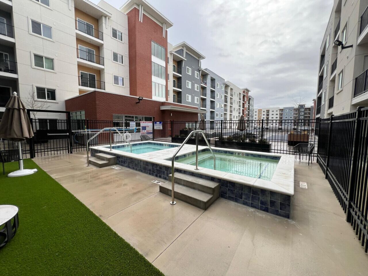 A swimming pool with steps leading into it, surrounded by a fence, and adjacent to a building with multiple balconies.