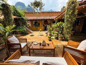 A patio with wooden furniture and potted plants, a yellow building with a tiled roof, and a mountainous backdrop.
