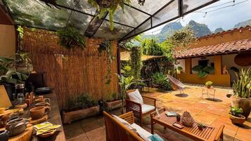 Un patio techado al aire libre con mobiliario de madera, plantas en macetas y vistas a un paisaje montañoso.