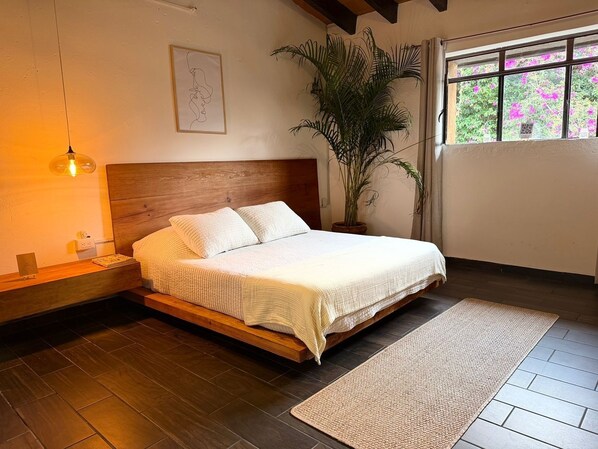 Une chambre à coucher avec un lit en bois, une table de chevet, une plante en pot et une fenêtre donnant sur un paysage verdoyant.