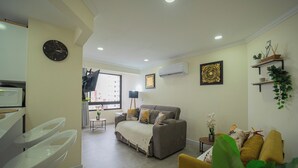 Uma sala de estar com um sofá, uma poltrona amarela, uma pequena mesa com uma planta, um relógio na parede e um aparelho de ar condicionado.