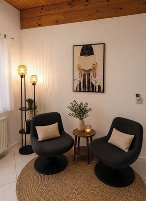 A room with two black armchairs, a round woven rug, a small table with a plant, a floor lamp, and a framed artwork on the wall.