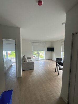 A spacious room with a large window, a flat-screen TV mounted on the wall, and a wooden floor. - Hotel W Des Cantons (Weedon)