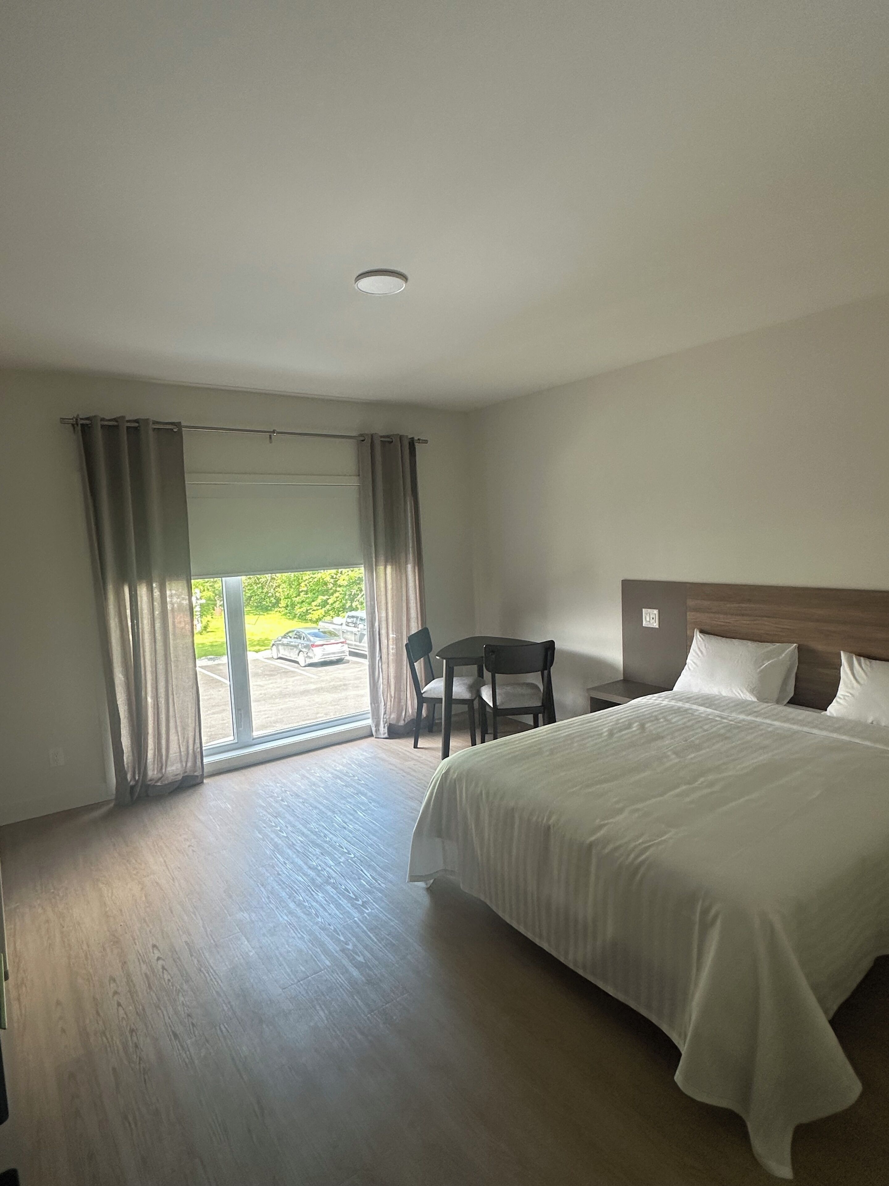 Habitación de hotel con una cama grande, un escritorio con dos sillas, vista a través de un ventanal y piso de madera.