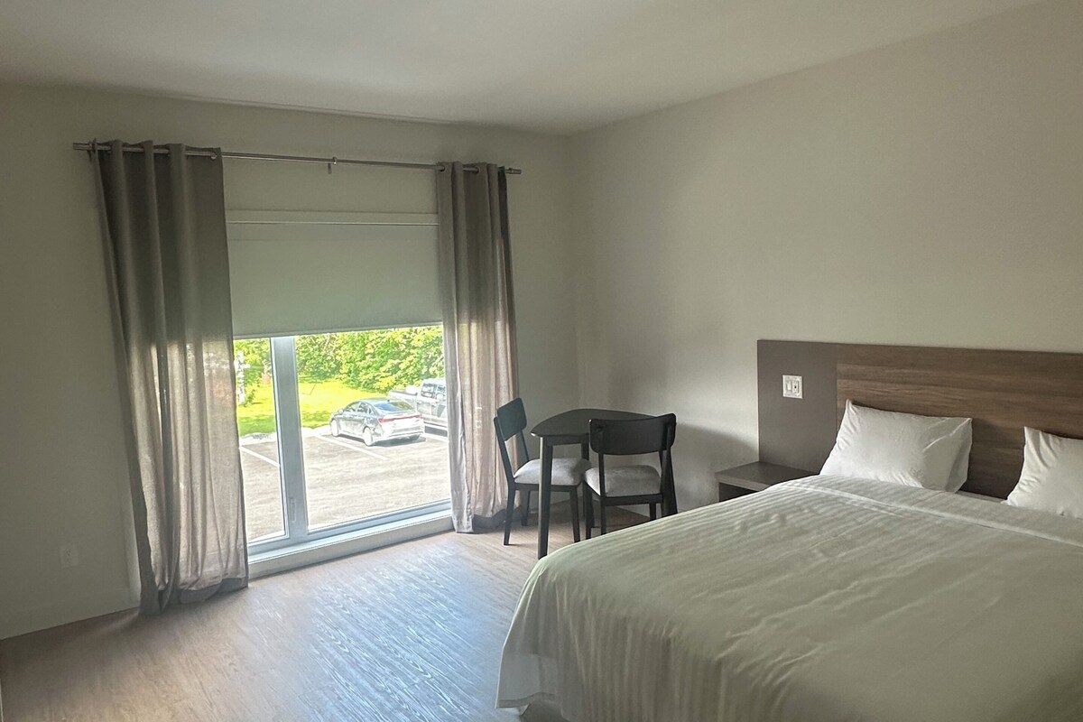 A hotel room with a large bed, a desk with two chairs, a view through a glass door, and a wooden floor.