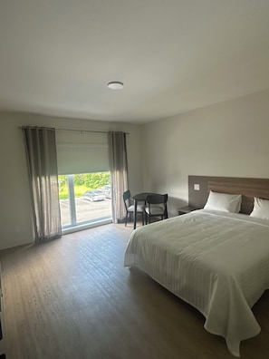 A hotel room with a large bed, a desk with two chairs, a view through a glass door, and a wooden floor. - Hotel W Des Cantons (Weedon)