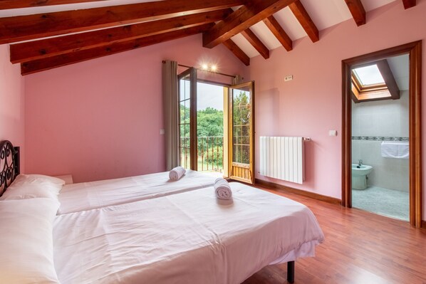 Un dormitorio con dos camas, vigas de madera, paredes rosas, un tragaluz y un baño visible a través de una puerta abierta.