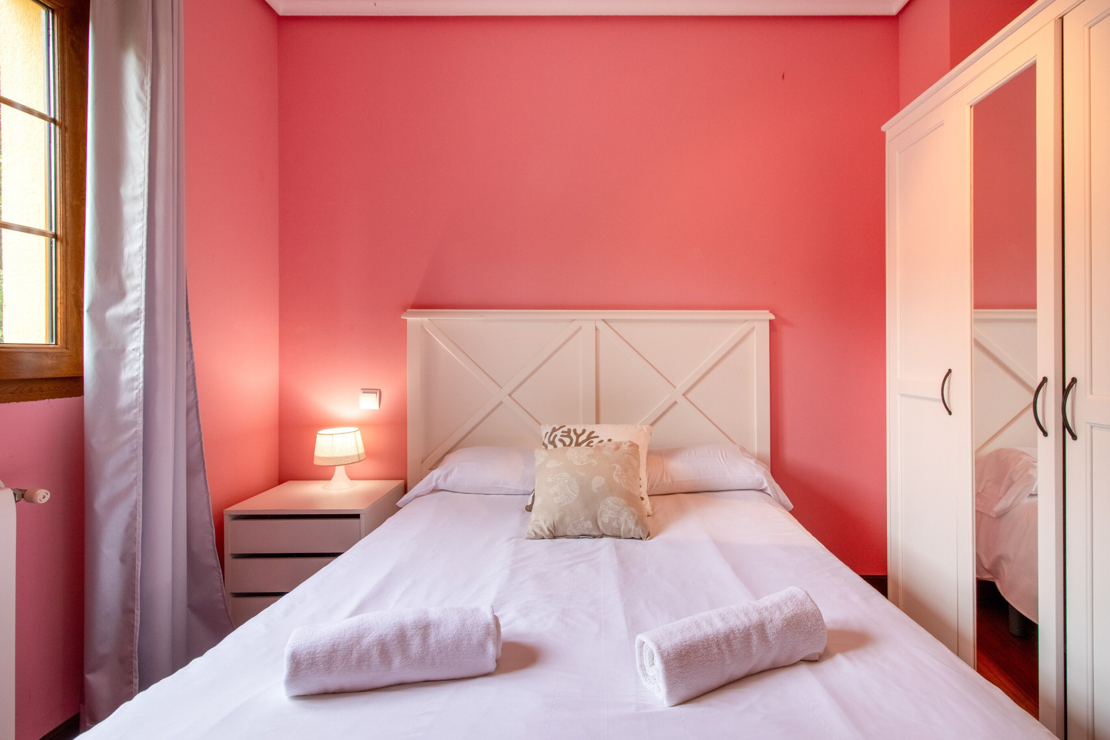 A bedroom with a bed, a nightstand, a lamp, a wardrobe, and a window with curtains.