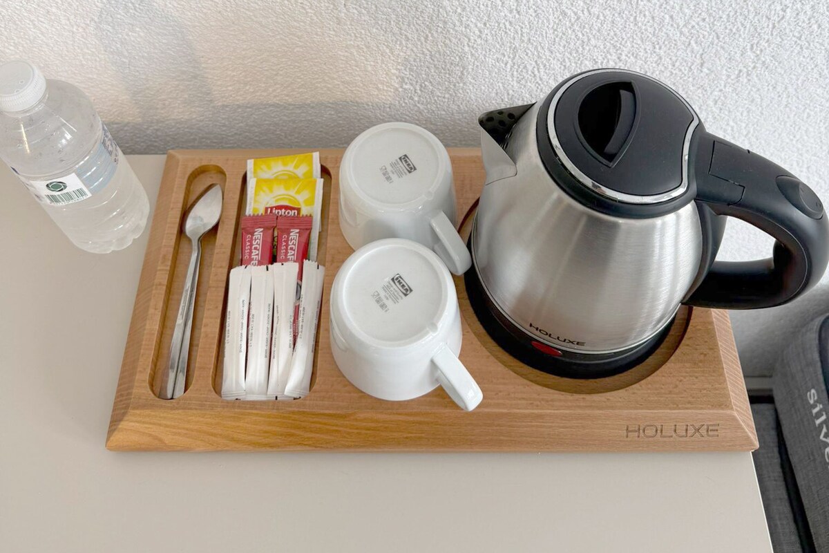 Una bandeja de madera con un hervidor, dos tazas blancas, una cuchara, una botella de agua y algunos sobres de café.