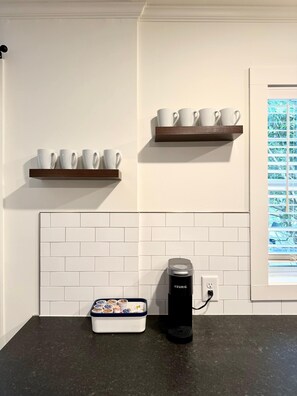 A kitchen with white tiled backsplash, two wooden shelves with white cups, a Keurig coffee maker, and a tray with food containers. - Charming Mid-Century Retreat | Pets Welcome (Raleigh)