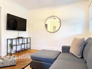 A living room with a grey sofa, a black TV stand with a plant, a round mirror, and a wooden floor. - Charming Mid-Century Retreat | Pets Welcome (Raleigh)