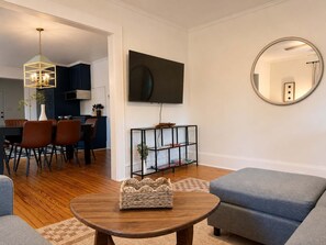 A modern living room with a wooden coffee table, grey sofas, a woven rug, a black TV, and a kitchen in the background. - Charming Mid-Century Retreat | Pets Welcome (Raleigh)