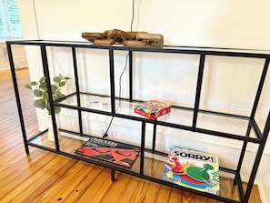 A black metal shelf unit with two board games, a potted plant, and a wooden sculpture. - Charming Mid-Century Retreat | Pets Welcome (Raleigh)