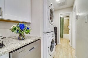 Un área de lavandería con lavadoras y secadoras apiladas, una encimera con un jarrón de flores y un pasillo que conduce a un baño.