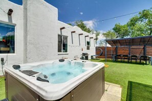 Un jacuzzi en un patio trasero con un edificio blanco, una terraza de madera y una zona de estar cubierta.