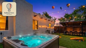 Un jacuzzi en un patio trasero con luces colgantes, un comedor y vista al cielo con globos aerostáticos.