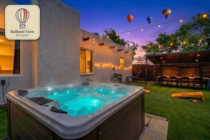 A hot tub in a backyard with string lights, a dining area, and a view of the sky with hot air balloons.