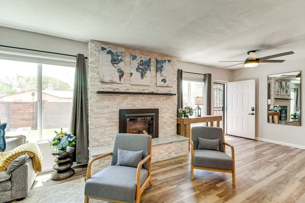 A living room with a fireplace, two grey armchairs, a wooden coffee table, and a large window.
