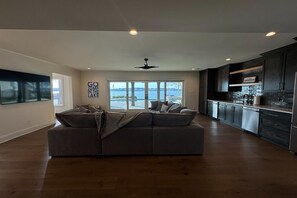 Un vaste espace de vie comprenant un grand canapé d’angle gris, un ventilateur de plafond et une vue sur un lac, visible à travers de grandes fenêtres.