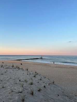 Ein Strand mit einer hölzernen Pier, die sich ins Wasser erstreckt, einem klaren Himmel und wenig Vegetation im Sand.