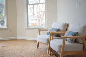 Ein Zimmer mit zwei hellen Stühlen mit blauen Kissen, einem beigen Teppich und einem großen Fenster mit Blick auf kahle Bäume.