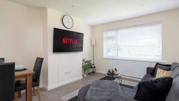 A living room with a flat-screen TV mounted on the wall, a dining area with a table and chairs, a sofa, and a large window with blinds.