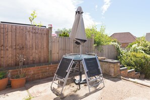 Eine Terrasse mit einem Metalltisch und -stühlen, einem grauen Sonnenschirm und einem Holzzaun.