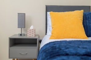 Ein Bett mit blauer Kopfteil, einem gelben Kissen und einem grauen Nachttisch mit Lampe und Taschentuchbox.