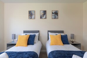 Ein Doppelzimmer mit zwei Betten, jeweils mit Kopfstütze, Nachttischen mit Lampen und drei abstrakten Bildern an der Wand.